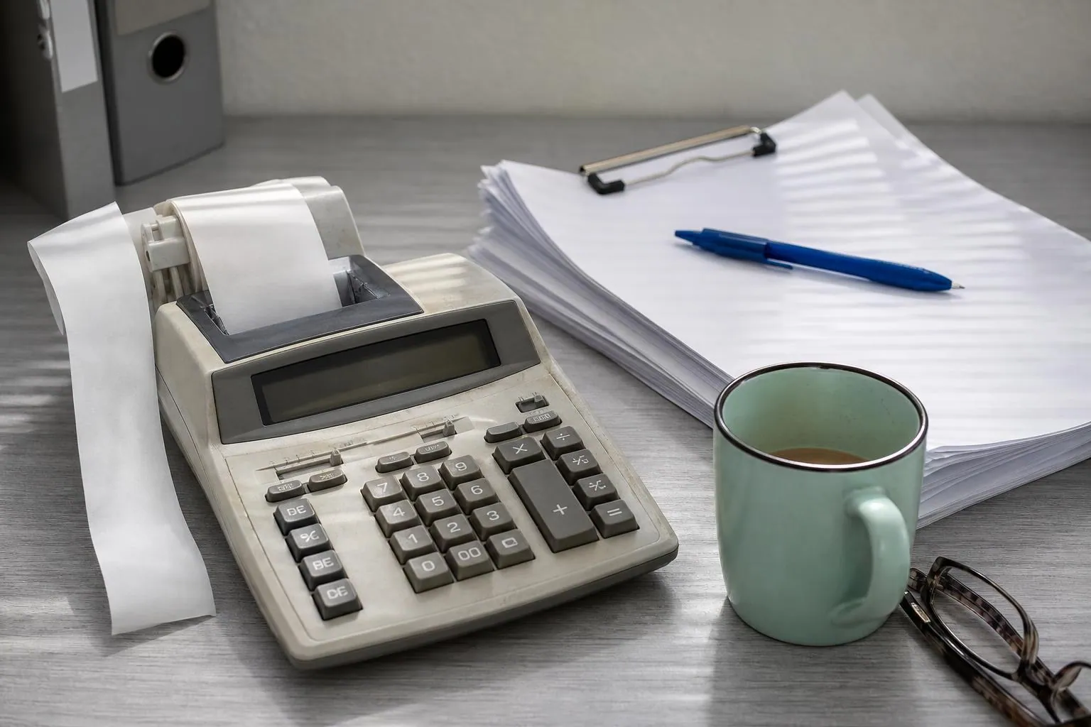 Calculatrice de bureau a rouleau de papier posee sur un plan de travail avec une liasse de factures papier et un mug en gres.