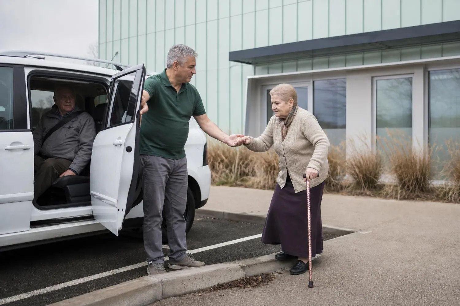Conducteur de taxi conventionne tenant la portiere arriere d un monospace blanc et aidant une patiente agee a monter, un second patient etant deja assis a l arriere, devant l entree d un centre de dialyse